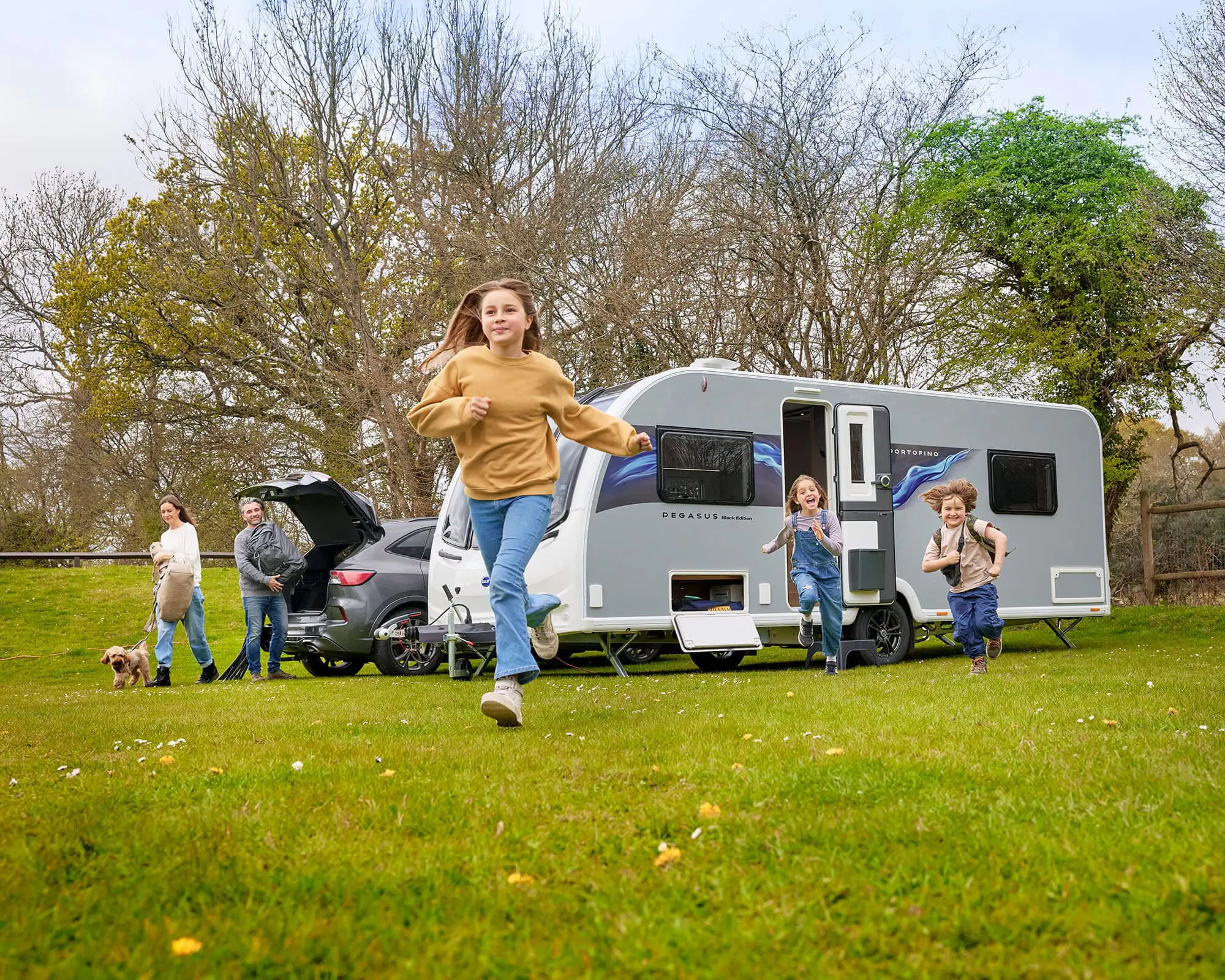 A family with children and a dog playing outside their Bailey caravan in a scenic green area.