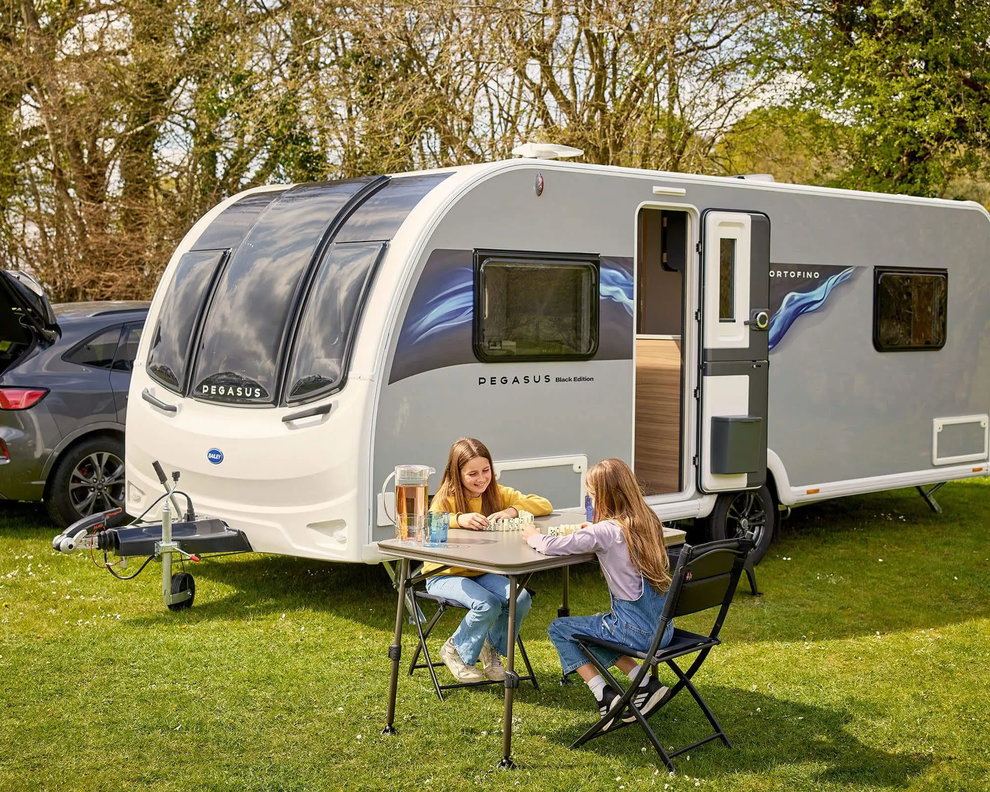Two children enjoying a meal outside a Bailey caravan, surrounded by greenery and parked vehicles.