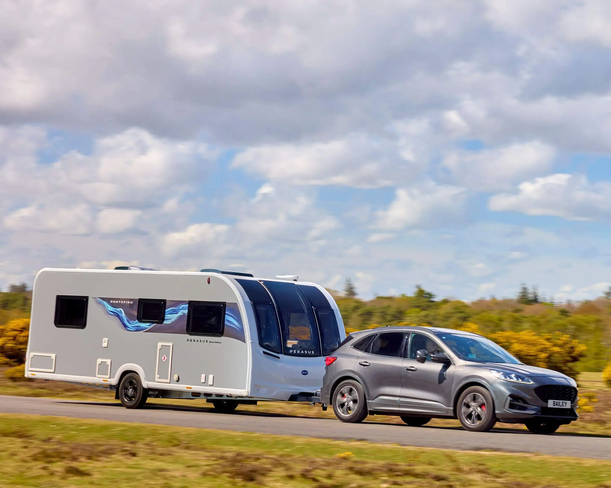 A silver vehicle towing a Bailey caravan on a picturesque landscape with blue skies and clouds.