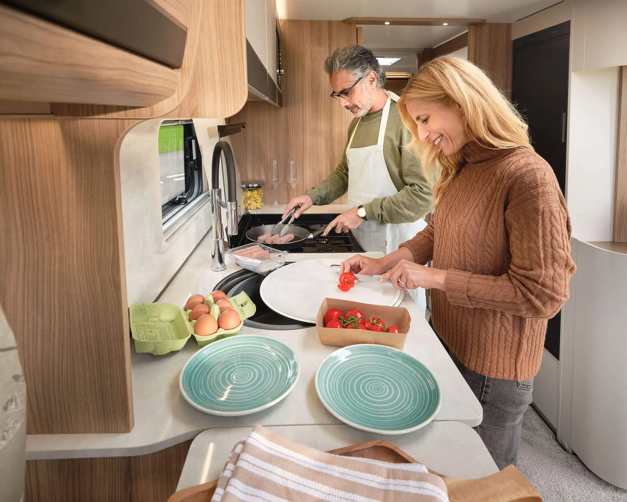 A couple preparing a meal in the kitchen of a Bailey caravan, featuring a cozy wooden interior and modern appliances.