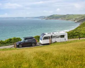 A Bailey caravan parked beside an SUV on a coastal road, overlooking the ocean and lush green hills.