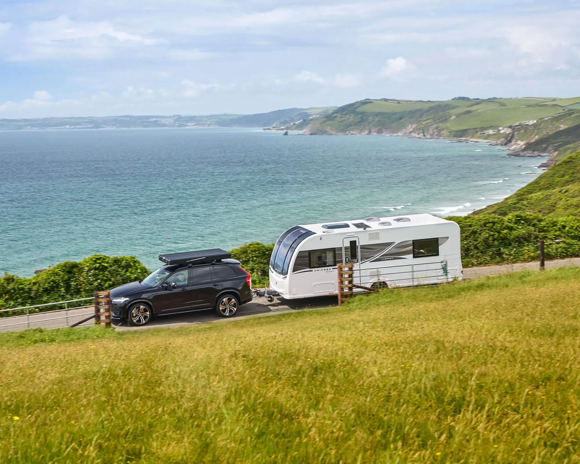 Scenic Coastal View with a Bailey Caravan and SUV A Bailey caravan parked beside an SUV on a coastal road, overlooking the ocean and lush green hills.