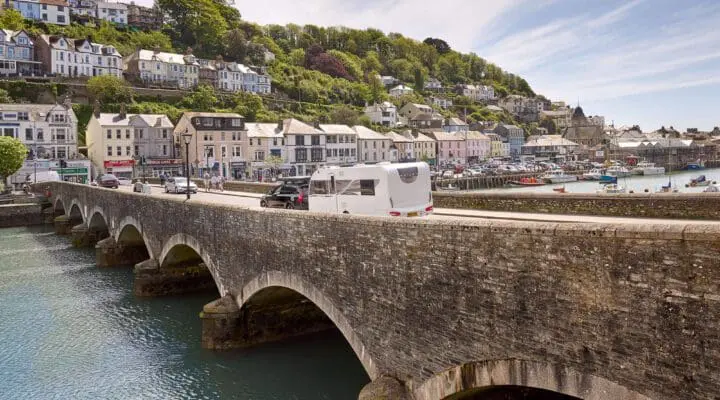A caravan driving over a stone bridge with a coastal village in the background, showcasing beautiful scenery.