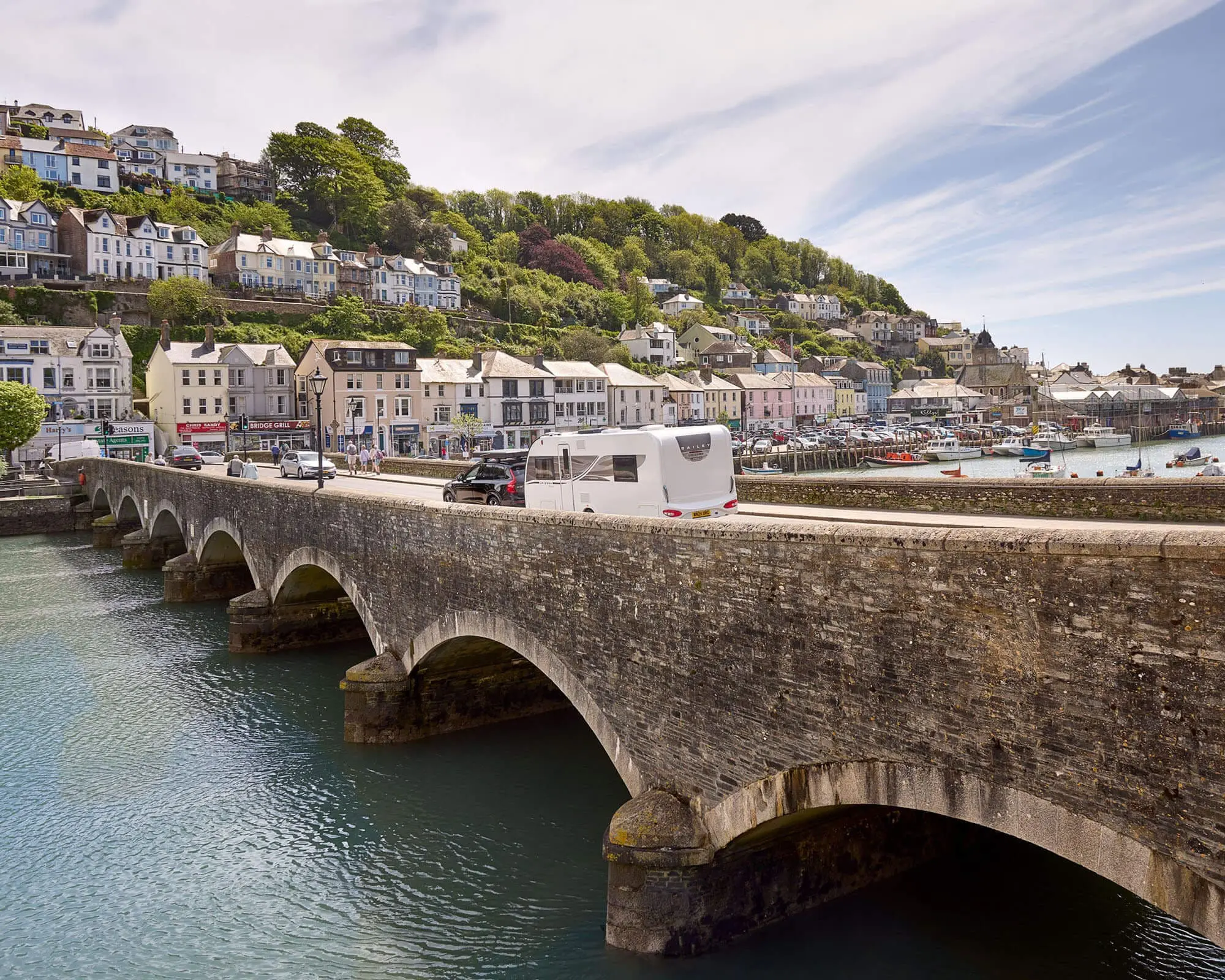 Scenic Bridge with Caravan Overlooking Coastal Village A caravan driving over a stone bridge with a coastal village in the background, showcasing beautiful scenery.
