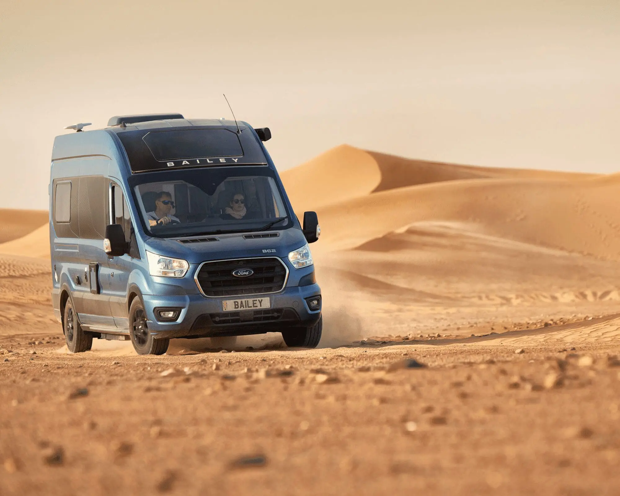 A blue Bailey Campervan driving through sandy desert dunes, showcasing durability and adventure travel potential.