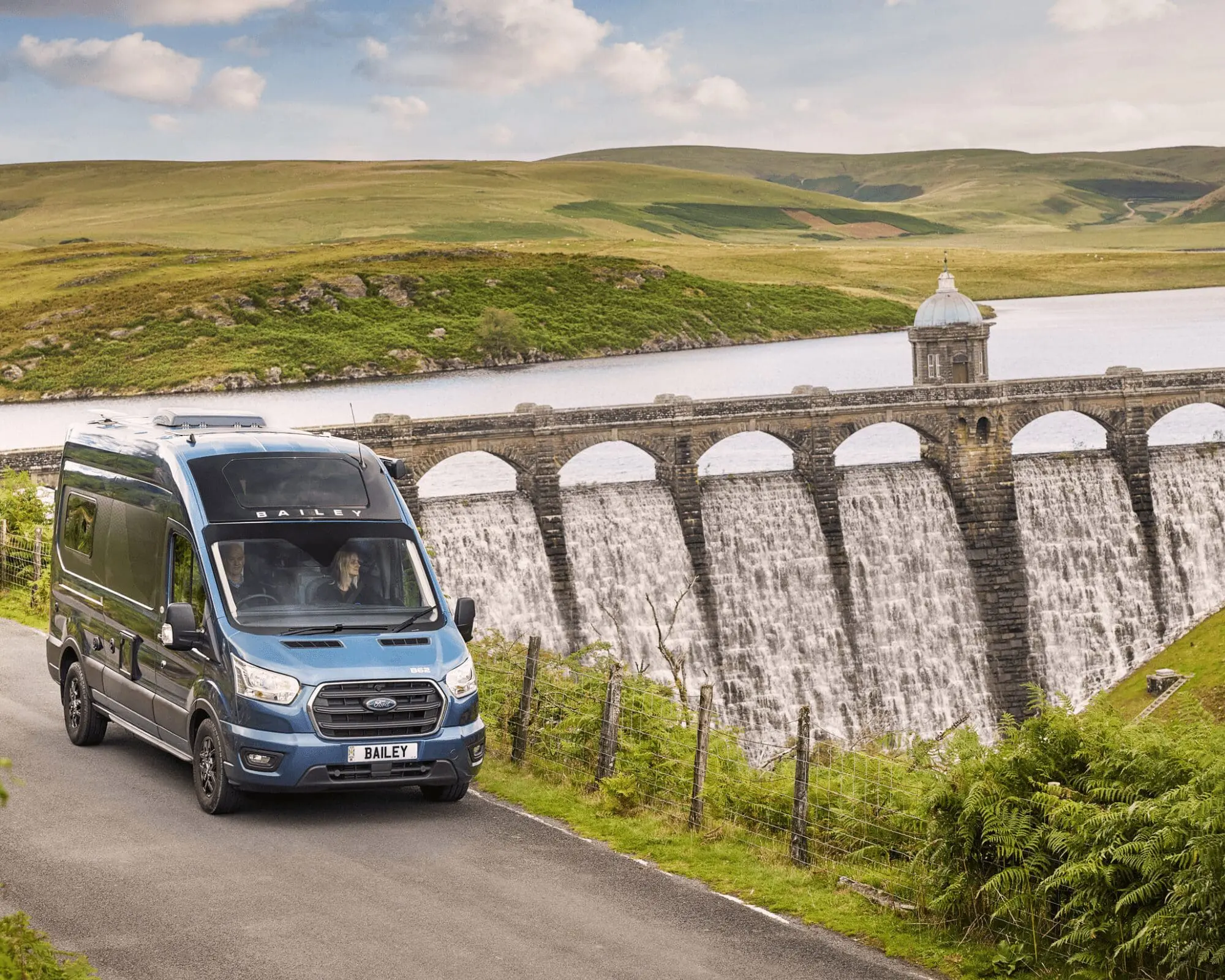 A blue Campervan driving on a road next to a large dam with cascading water, surrounded by green hills.