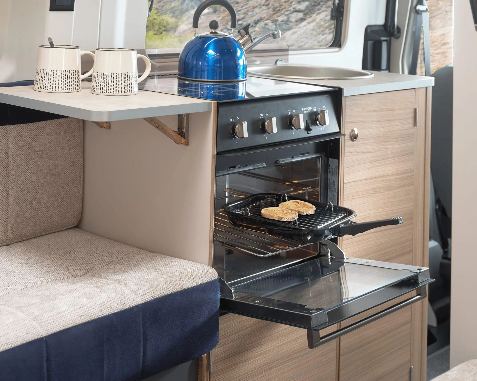 Interior of a caravan kitchen featuring an oven, a blue kettle, and a cozy seating area.