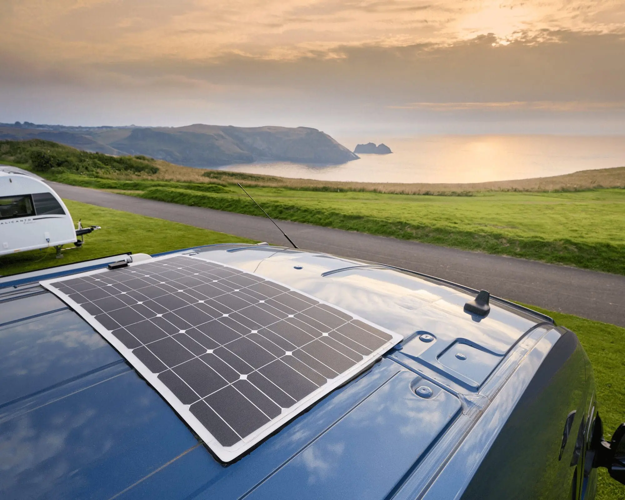 A solar panel installed on the roof of a campervan, overlooking a scenic coastal landscape at sunset.