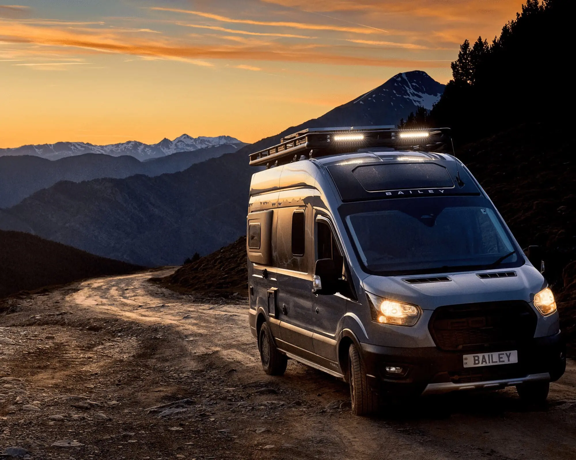 A grey campervan parked on a rocky road with mountains in the background during sunset.