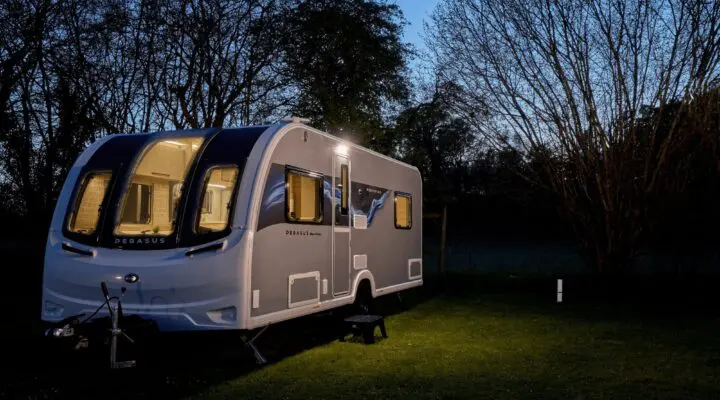 A sleek Bailey caravan illuminated at dusk, surrounded by trees in a serene setting.
