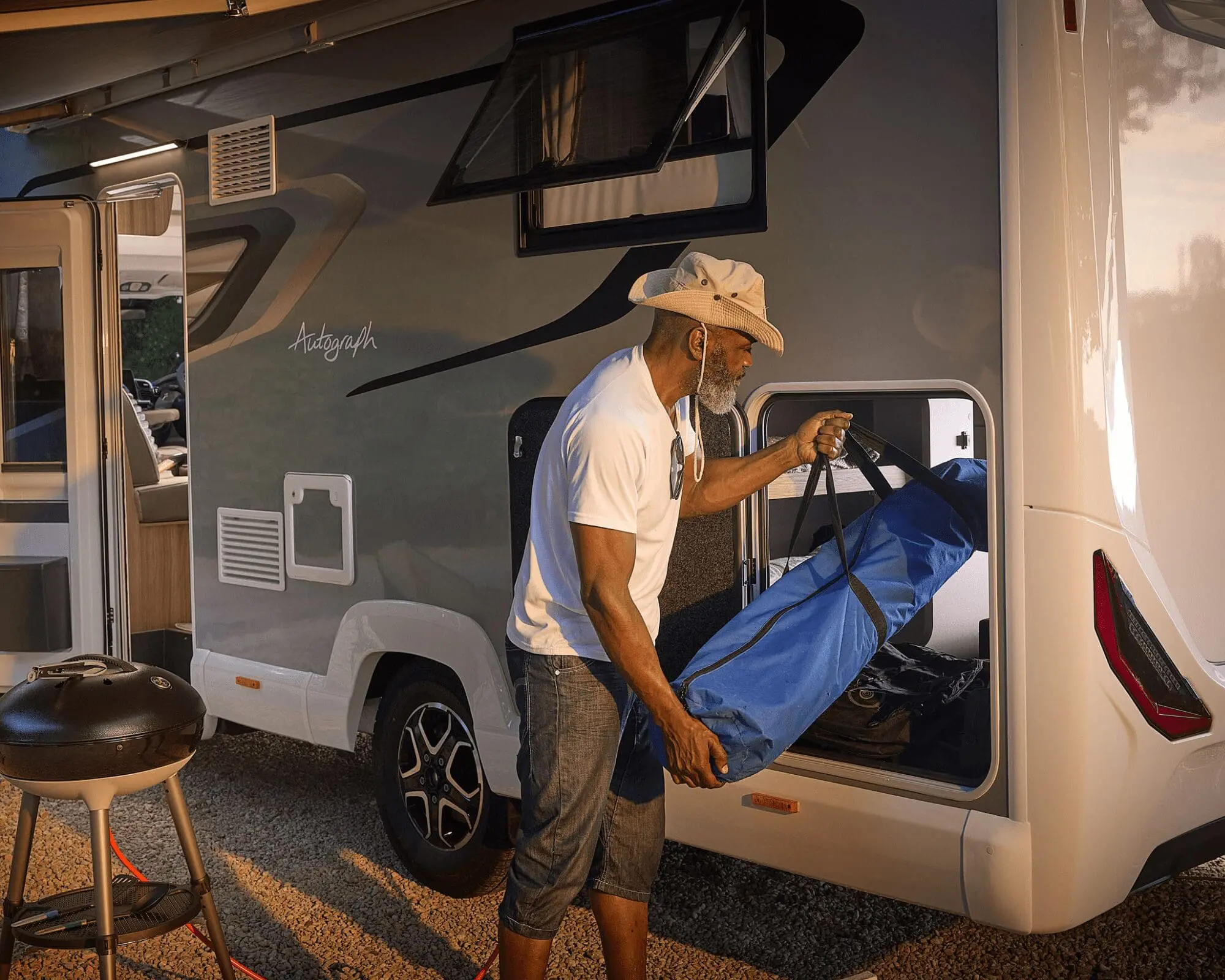 A man with a hat taking out a blue bag from a Bailey motorhome during sunset.