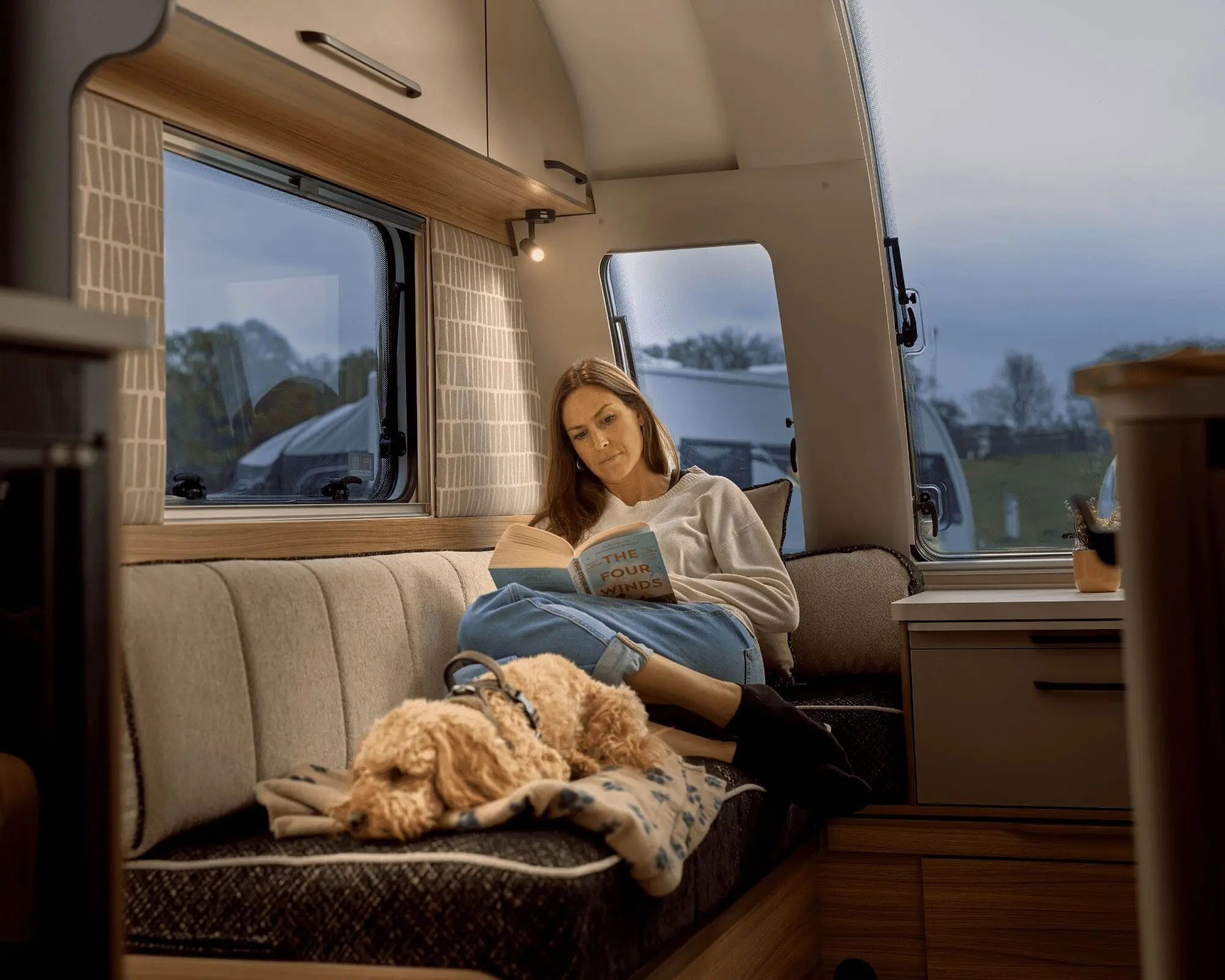 A woman sitting comfortably in a Bailey motorhome interior, reading a book with a dog on her lap, showcasing a warm and inviting space.