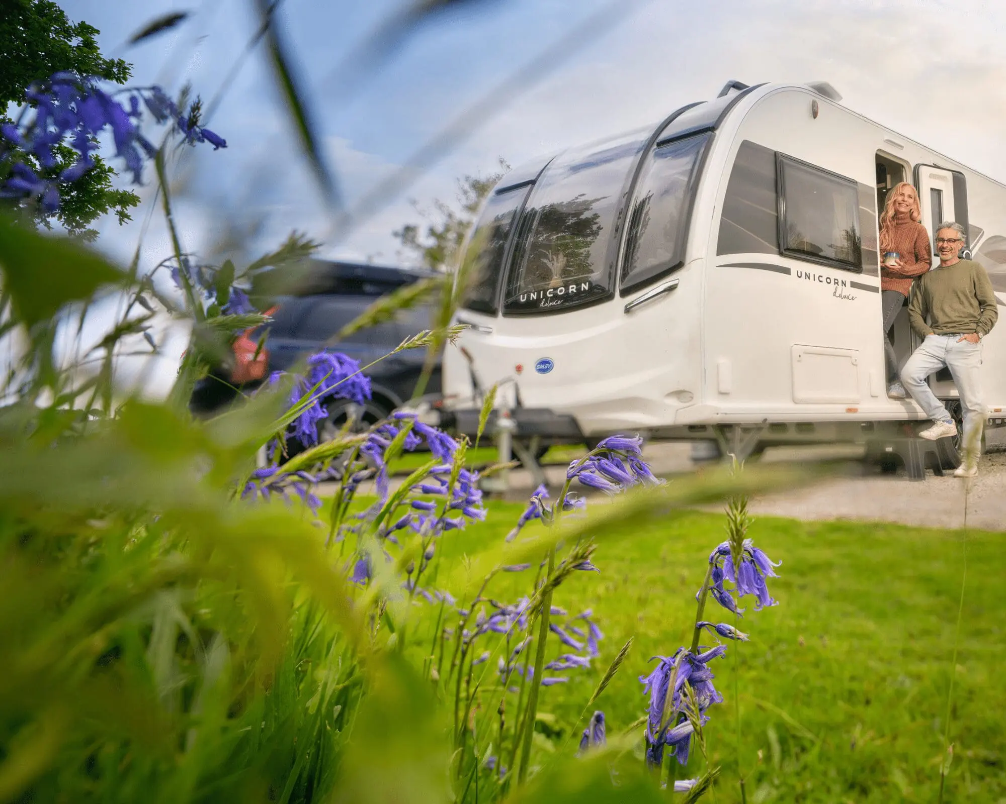 A Bailey caravan parked in a green outdoor environment surrounded by flowers, with people enjoying the view.