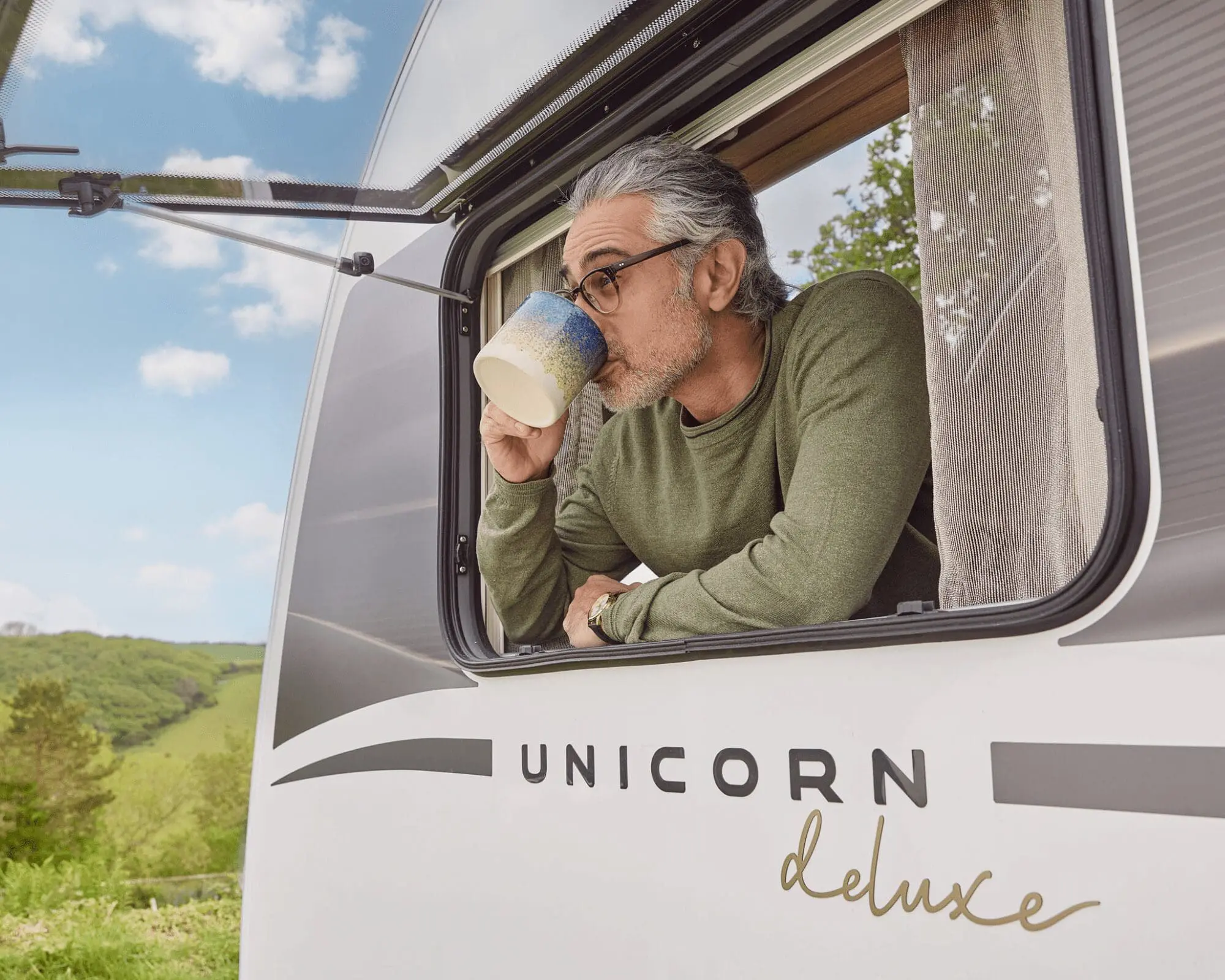 Man enjoying a drink from the window of a Bailey Unicorn Deluxe caravan surrounded by nature