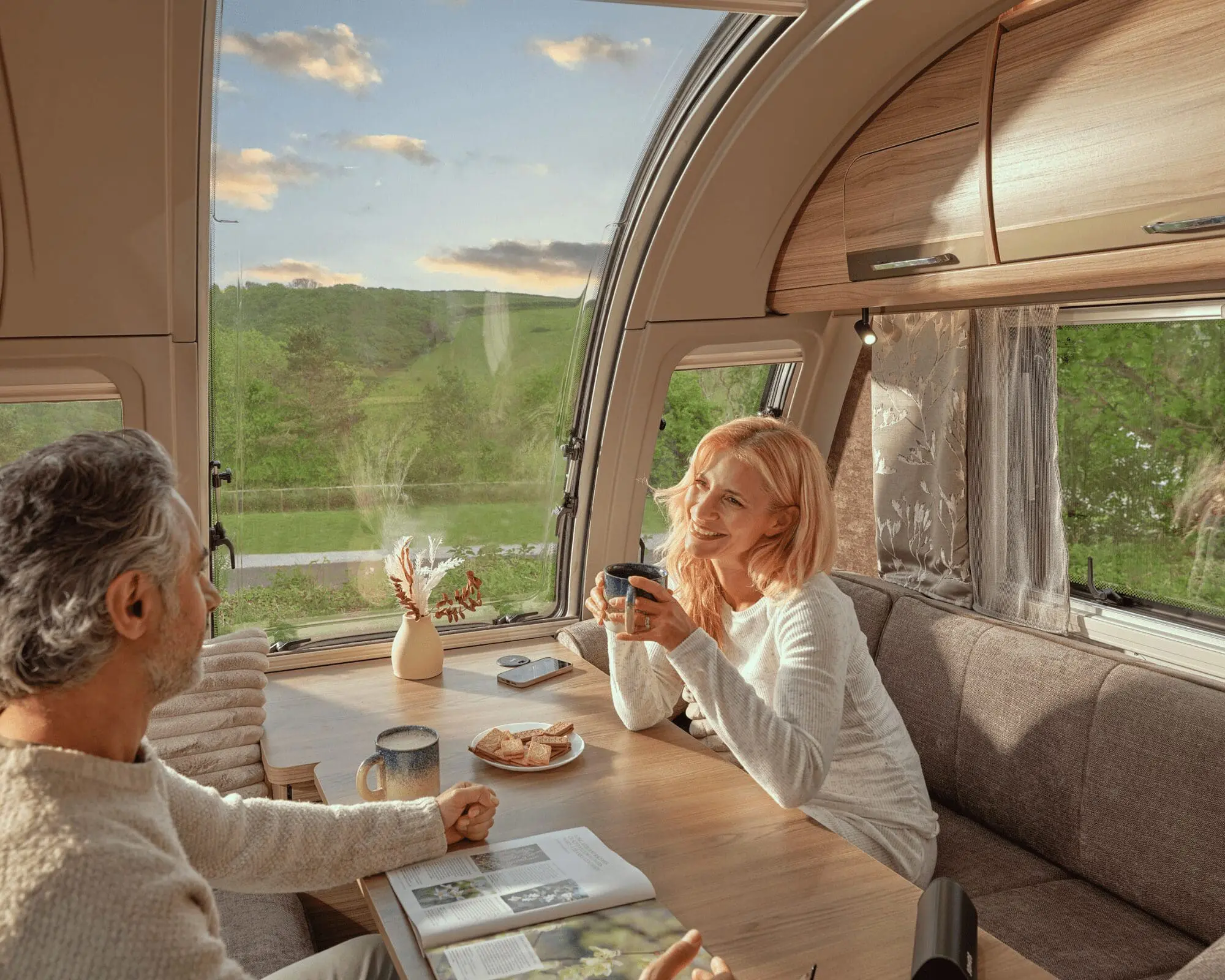Two people enjoying coffee inside a Bailey caravan, surrounded by modern decor and a scenic view through the window.