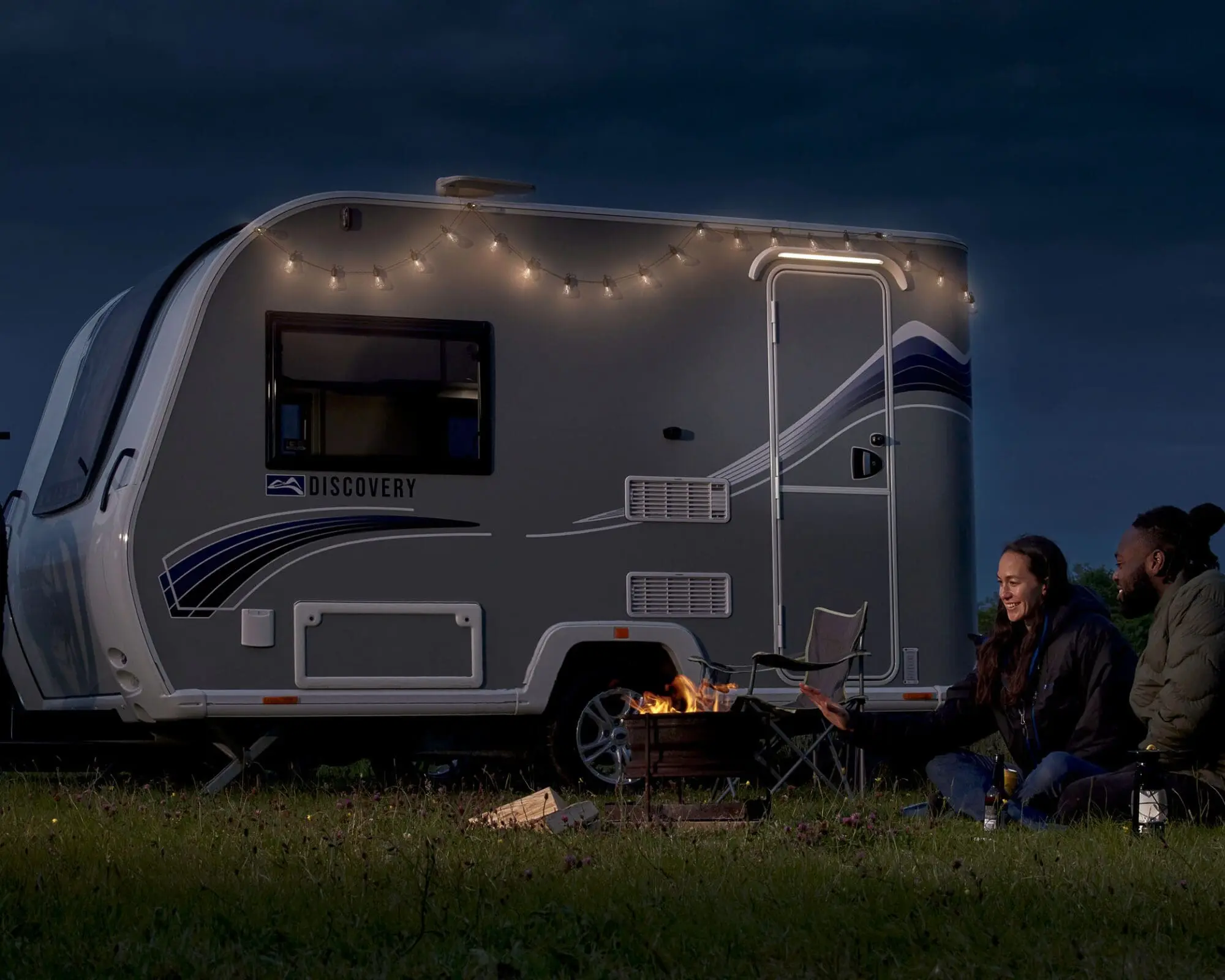 Cozy Evening by a Bailey Caravan with Fire Pit A Bailey caravan illuminated by string lights at dusk with two people enjoying a fire pit