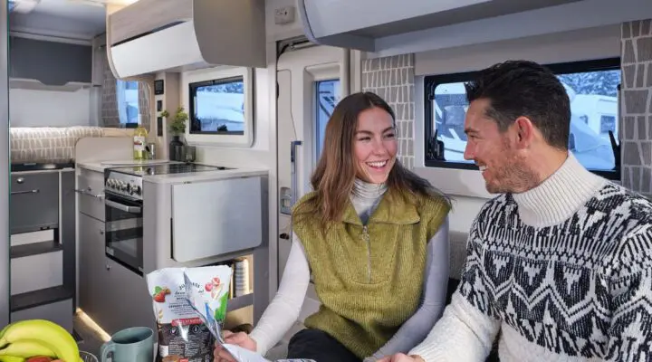 A couple sitting at a table in a modern caravan kitchen enjoying breakfast together, surrounded by fresh fruits and cooking utensils.