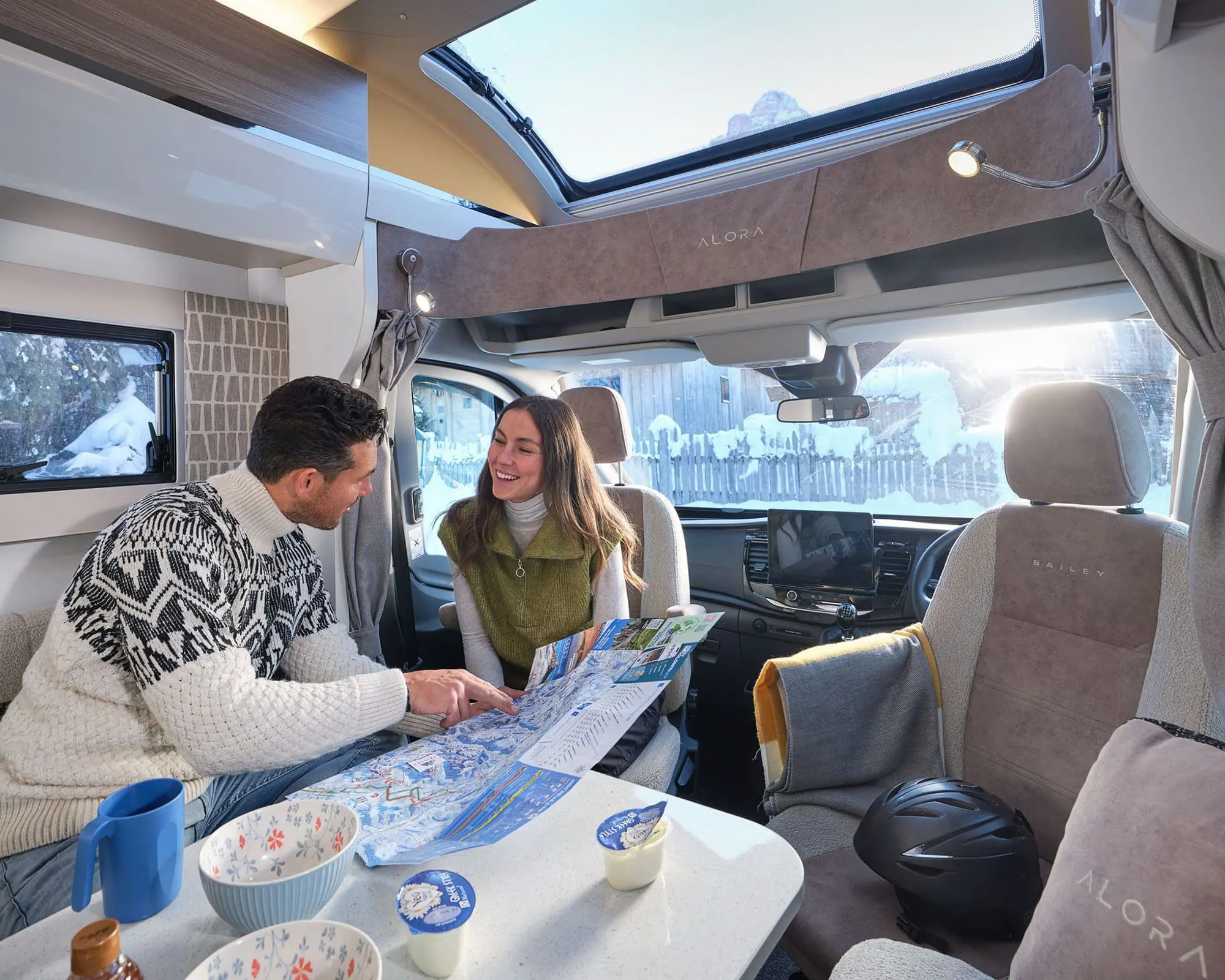 A couple examining a map while seated at a table inside a motorhome, with bright sunlight streaming through the windows.