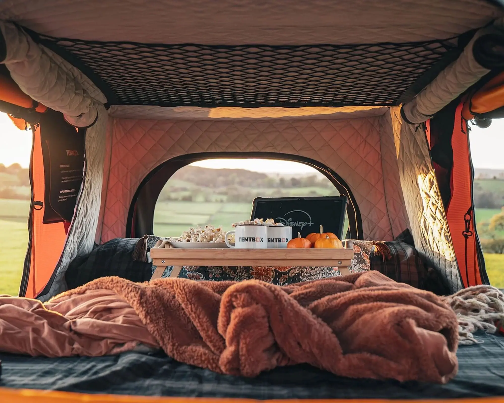 Interior view of a vehicle tent featuring a bed setup with a cozy blanket, a wooden table, and a beautiful natural landscape visible through the opening.