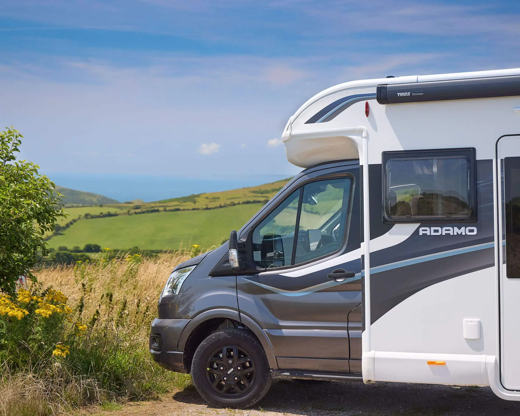 Side view of a Bailey motorhome parked in a beautiful rural setting with hills and a blue sky in the background.