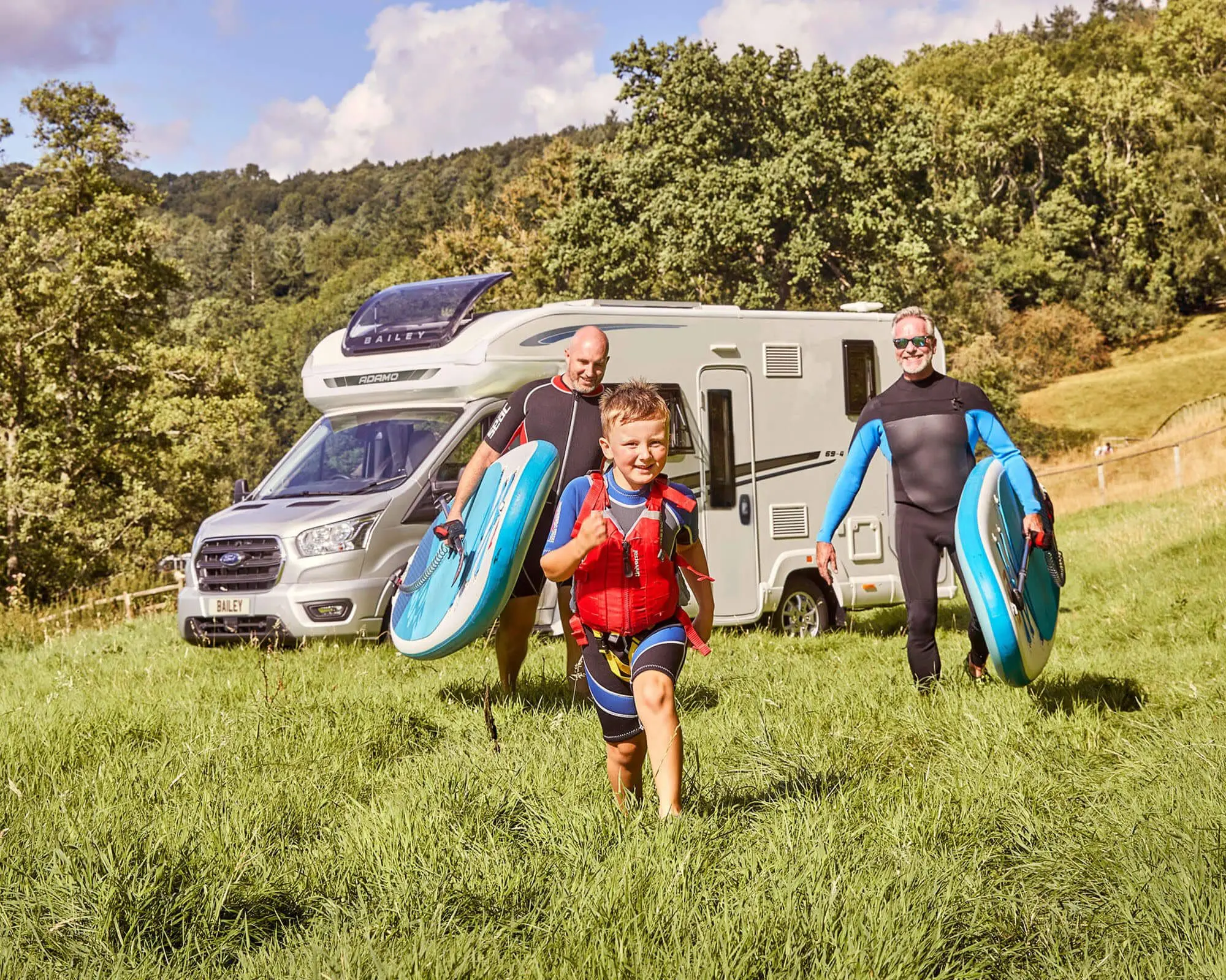 A child and two adults carrying surfboards while standing in a grassy field beside a camper van, surrounded by trees.