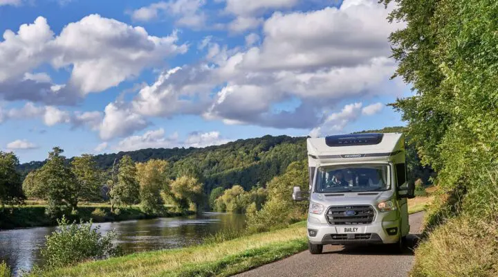 A Bailey motorhome parked alongside a scenic river with lush green hills and a blue sky filled with clouds.