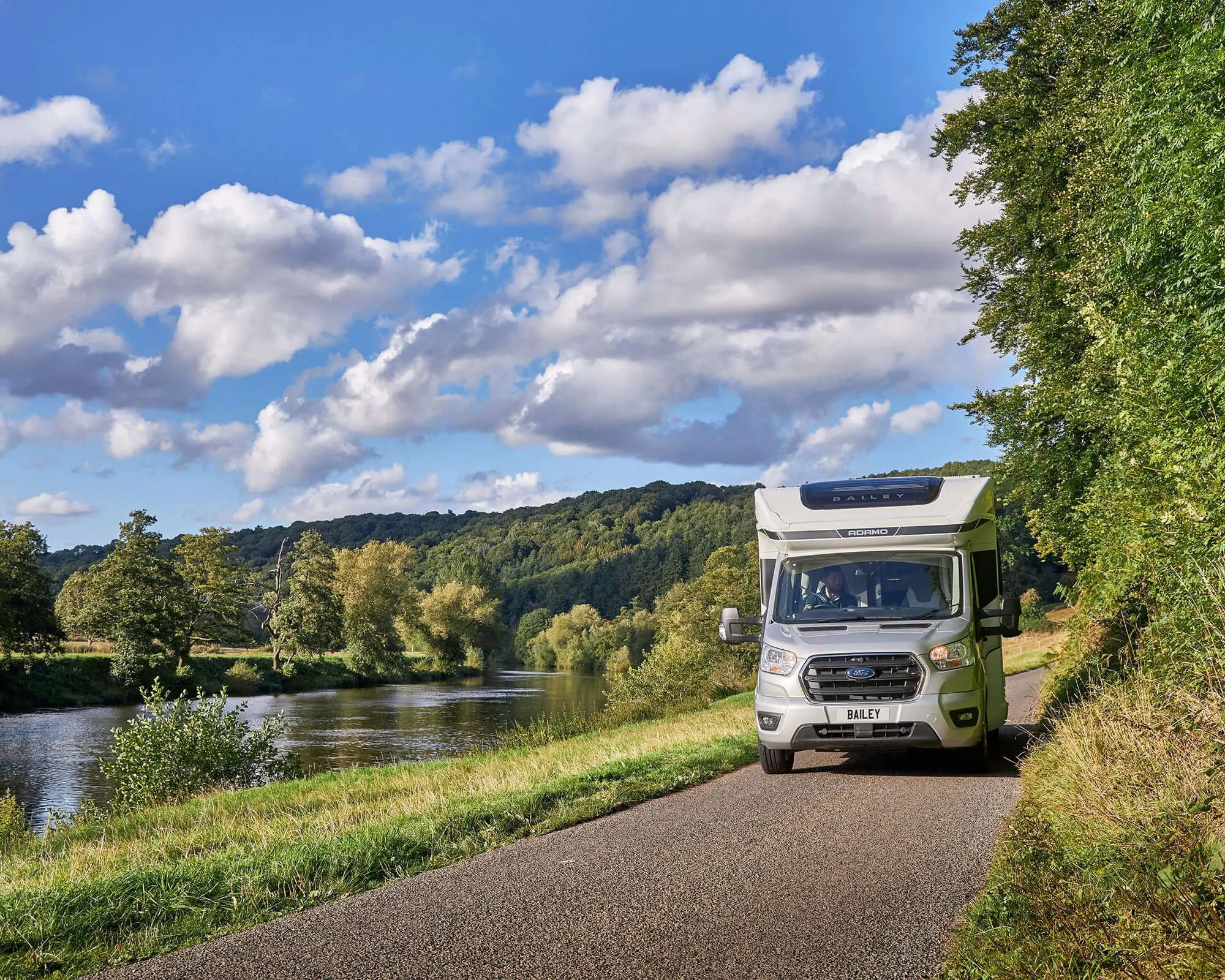 A Bailey motorhome parked alongside a scenic river with lush green hills and a blue sky filled with clouds.