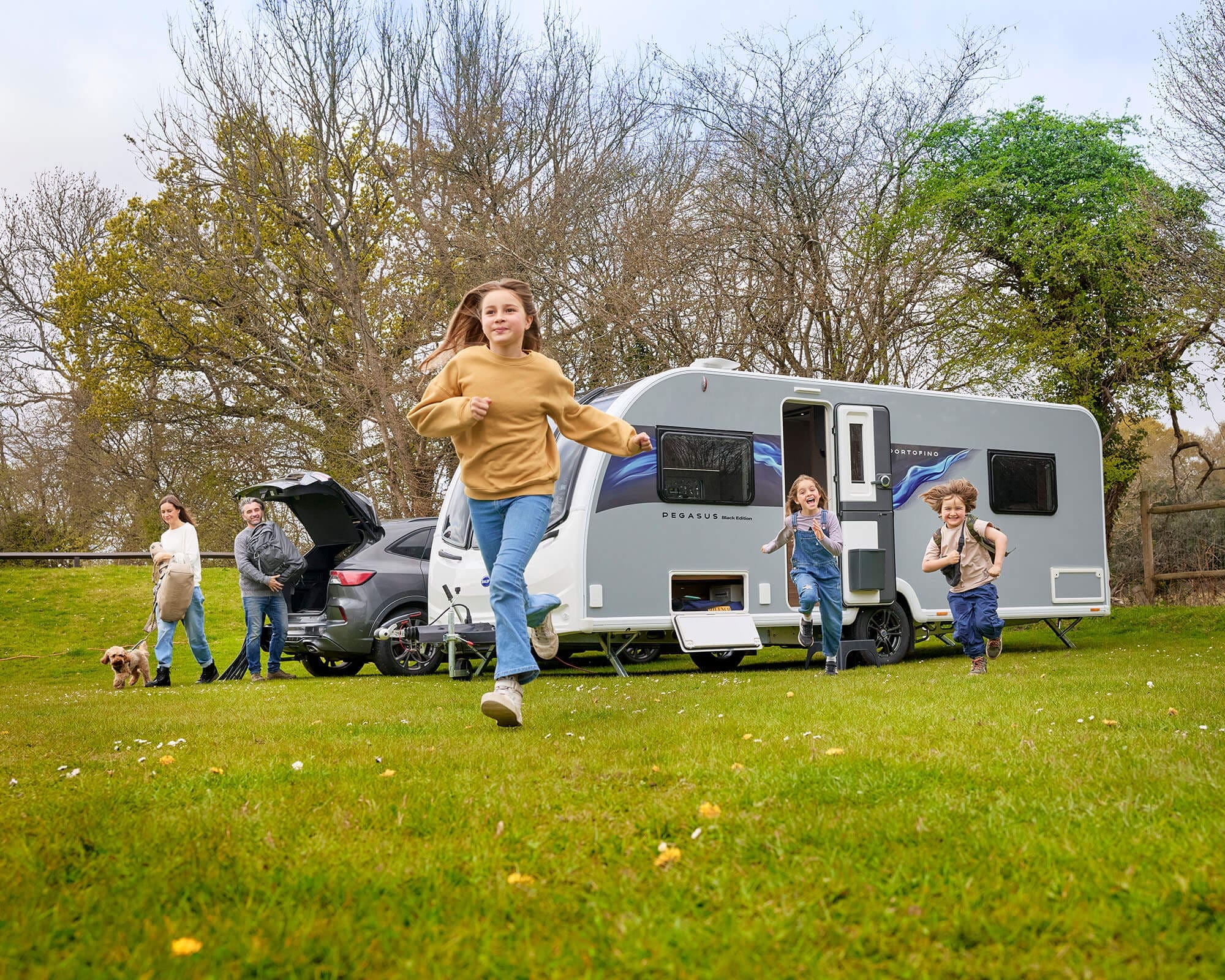 A family with children and a dog playing outside their Bailey caravan in a scenic green area.