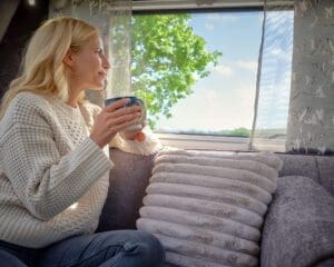 Woman enjoying a cup of coffee while looking out the window of a stylish caravan interior featuring cushions and natural light
