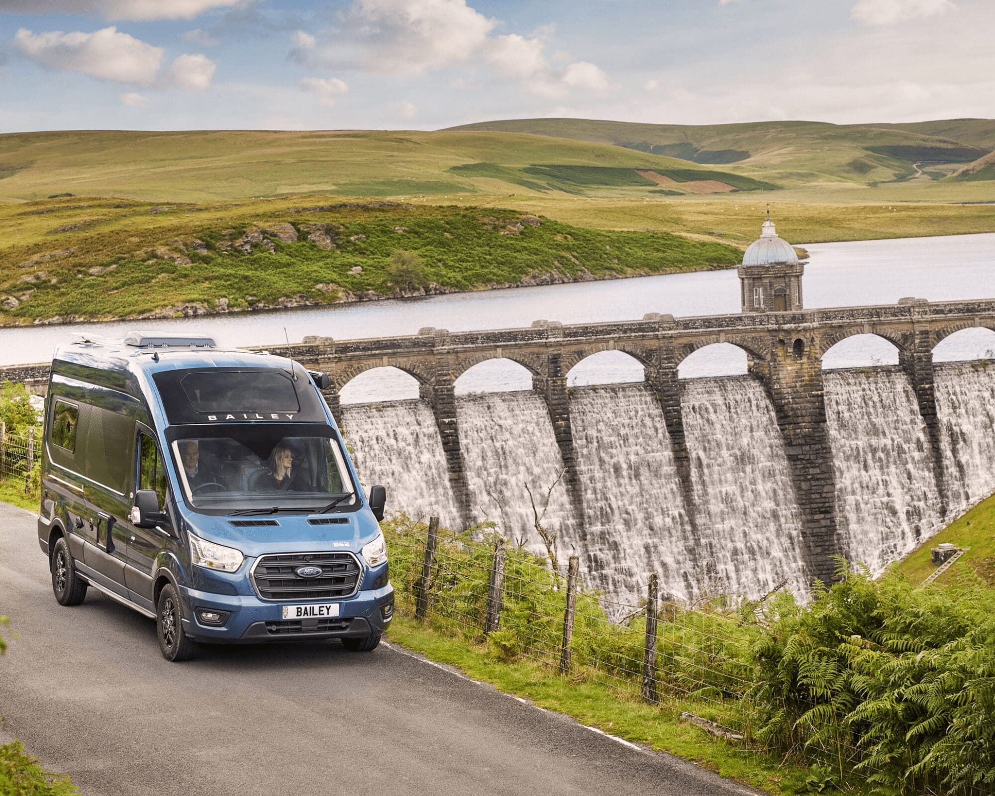 Blue Campervan in Scenic Landscape near Waterfall Dam A blue Campervan driving on a road next to a large dam with cascading water, surrounded by green hills.