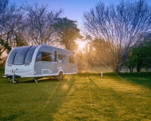 A sleek modern caravan parked in a lush green field during sunset, surrounded by trees.