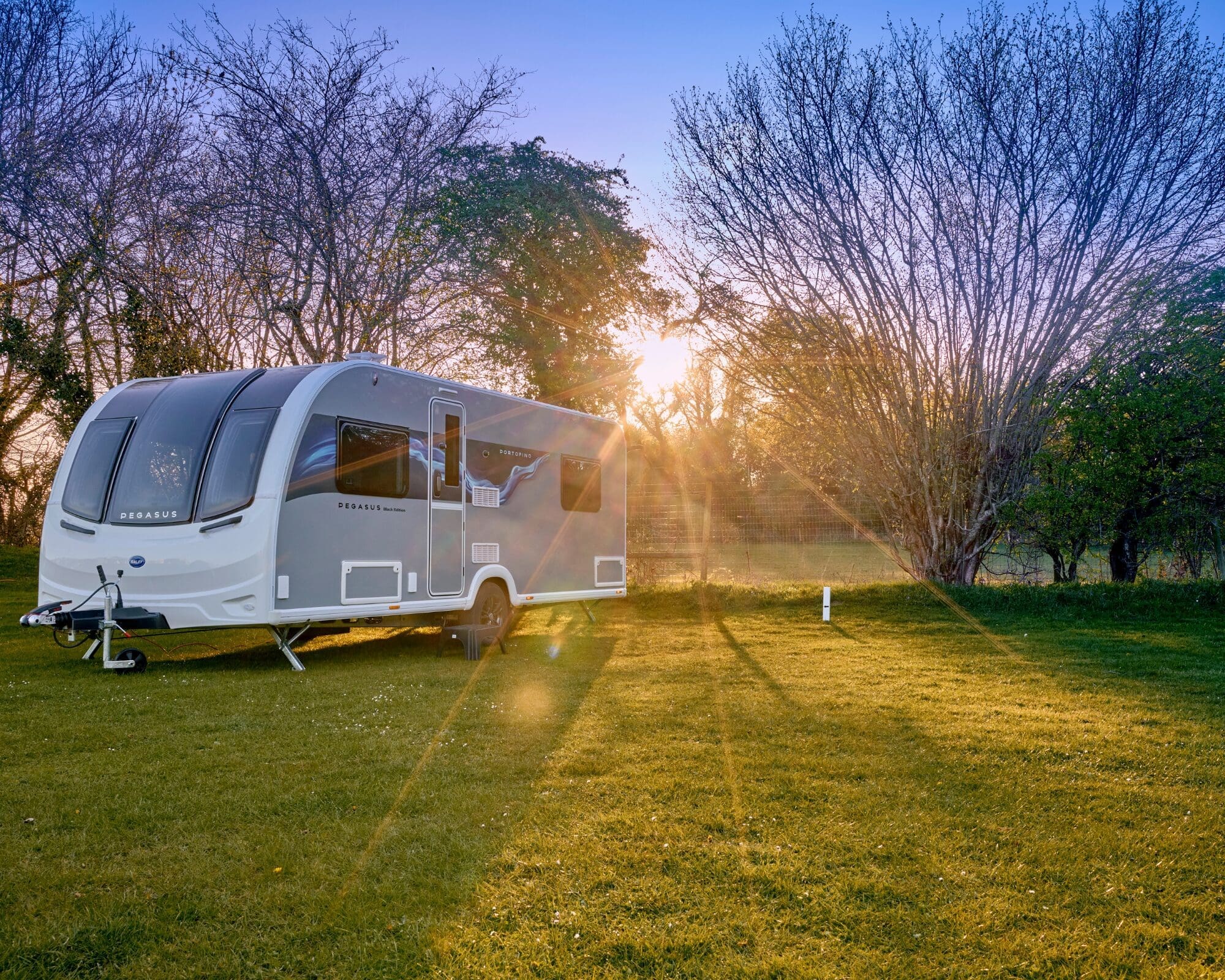 A sleek modern caravan parked in a lush green field during sunset, surrounded by trees.
