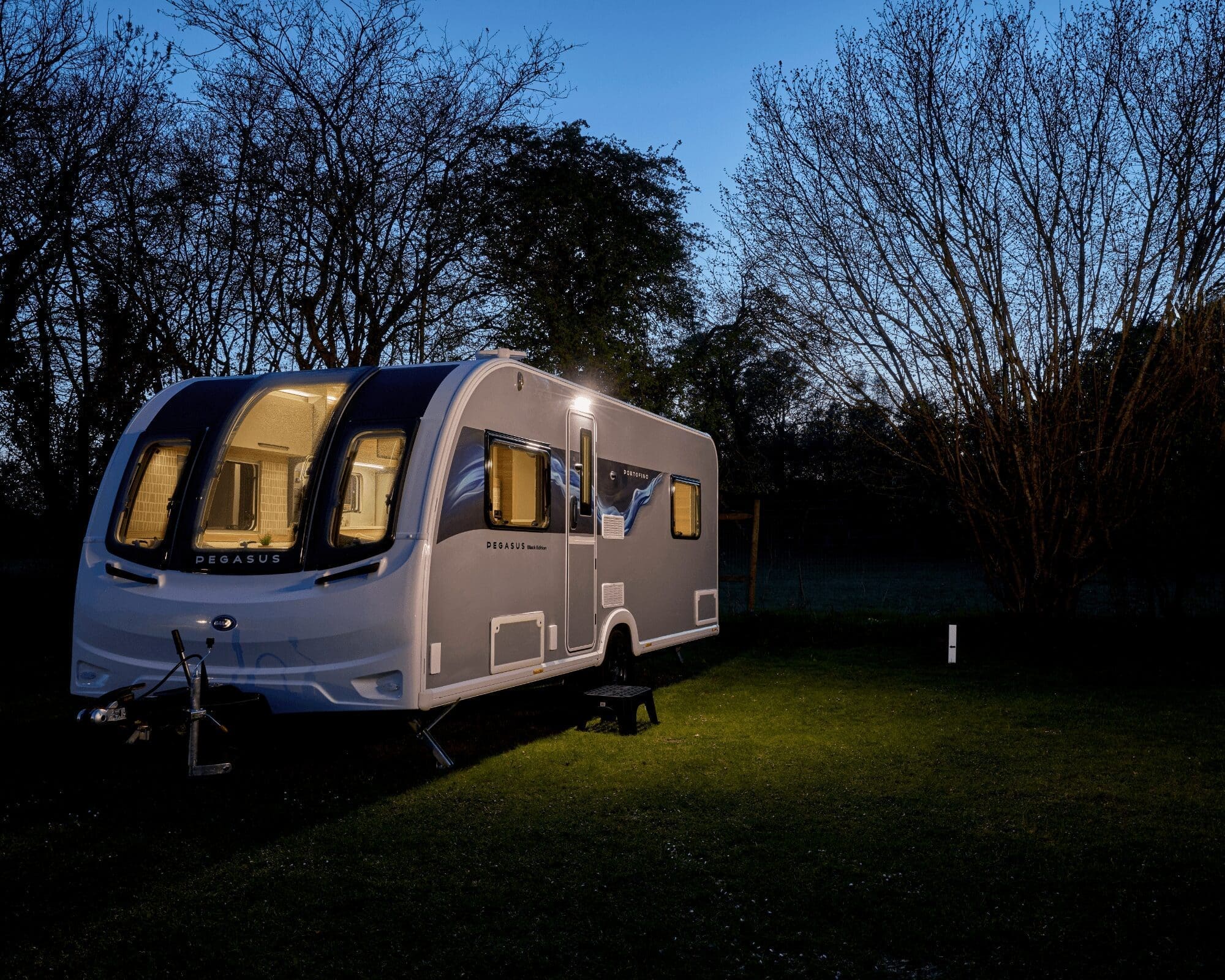 A sleek Bailey caravan illuminated at dusk, surrounded by trees in a serene setting.