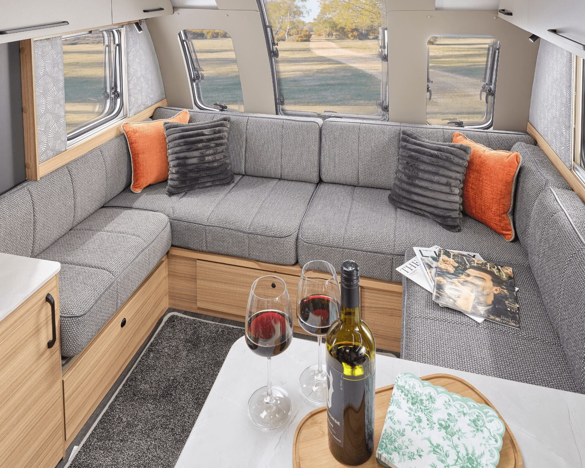 Cozy living area in a Bailey caravan featuring a grey upholstered couch, vibrant throw pillows, a dining table with wine glasses, and a scenic view outside.