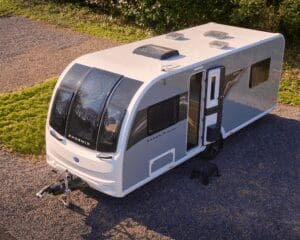 Aerial view of a stylish white caravan with large windows parked on gravel surrounded by greenery.