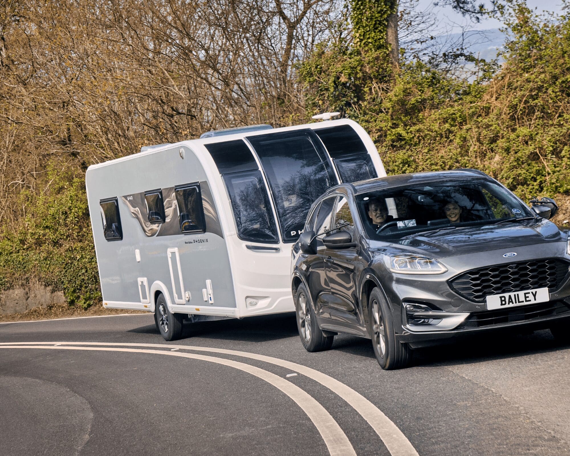 A modern Bailey caravan being towed by a sleek SUV along a curved road surrounded by trees, showcasing a perfect outdoor adventure setup.