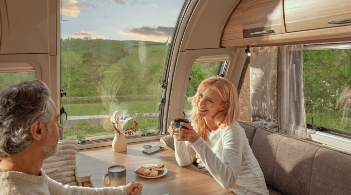 Two people enjoying coffee inside a Bailey caravan, surrounded by modern decor and a scenic view through the window.
