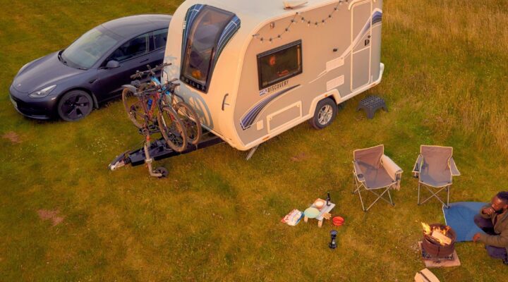 A Bailey caravan parked on grass next to a modern car, with outdoor camping chairs, a fire pit, and a relaxed camping setup.