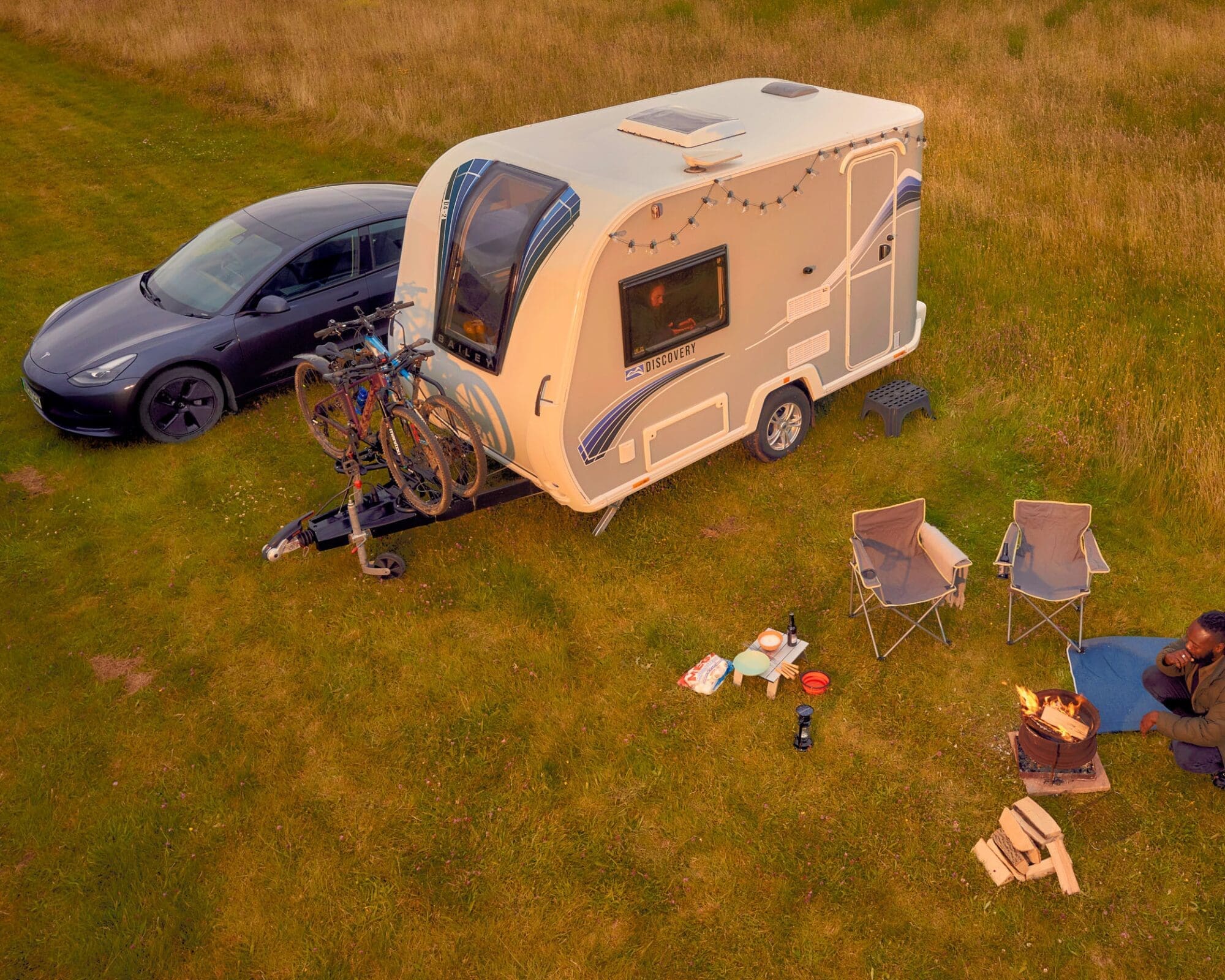 A Bailey caravan parked on grass next to a modern car, with outdoor camping chairs, a fire pit, and a relaxed camping setup.