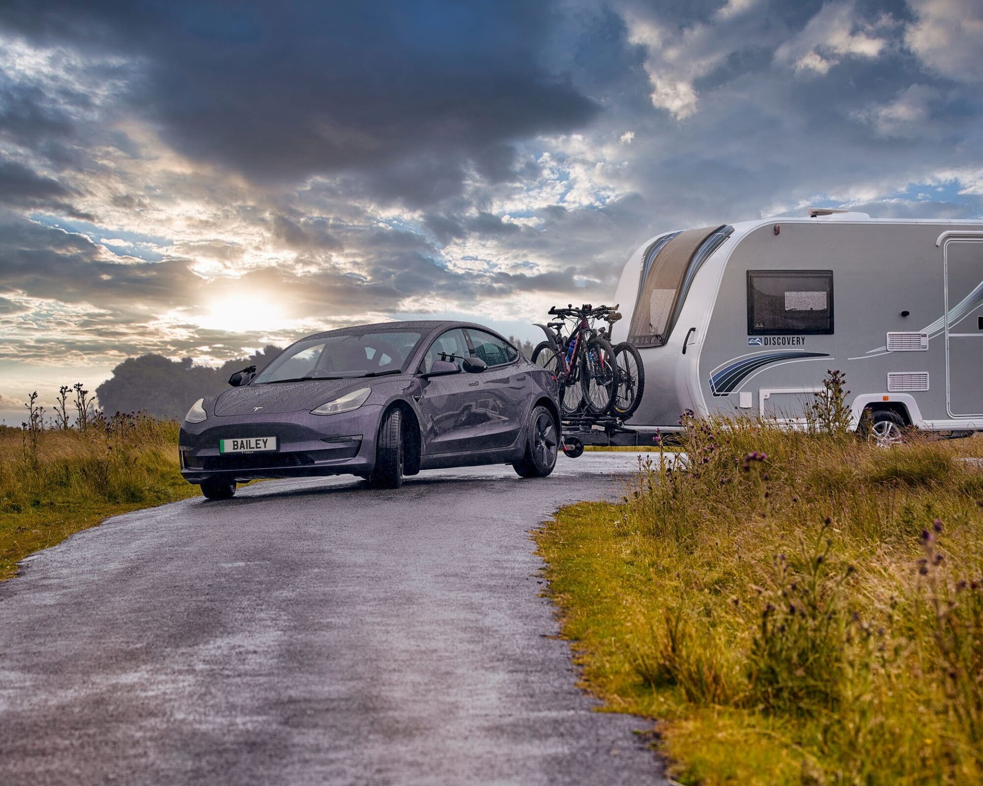 A stylish electric car towing a Bailey caravan on a scenic road during sunset, showcasing adventure and outdoor lifestyle.
