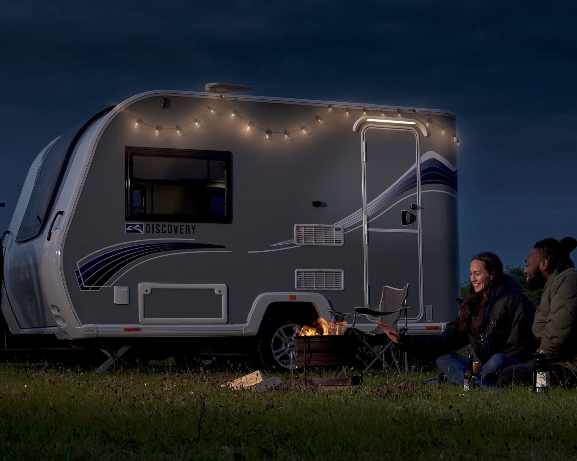 A Bailey caravan illuminated by string lights at dusk with two people enjoying a fire pit