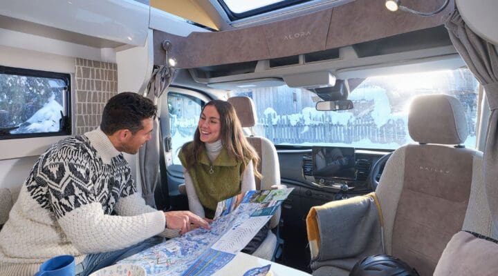 A couple examining a map while seated at a table inside a motorhome, with bright sunlight streaming through the windows.
