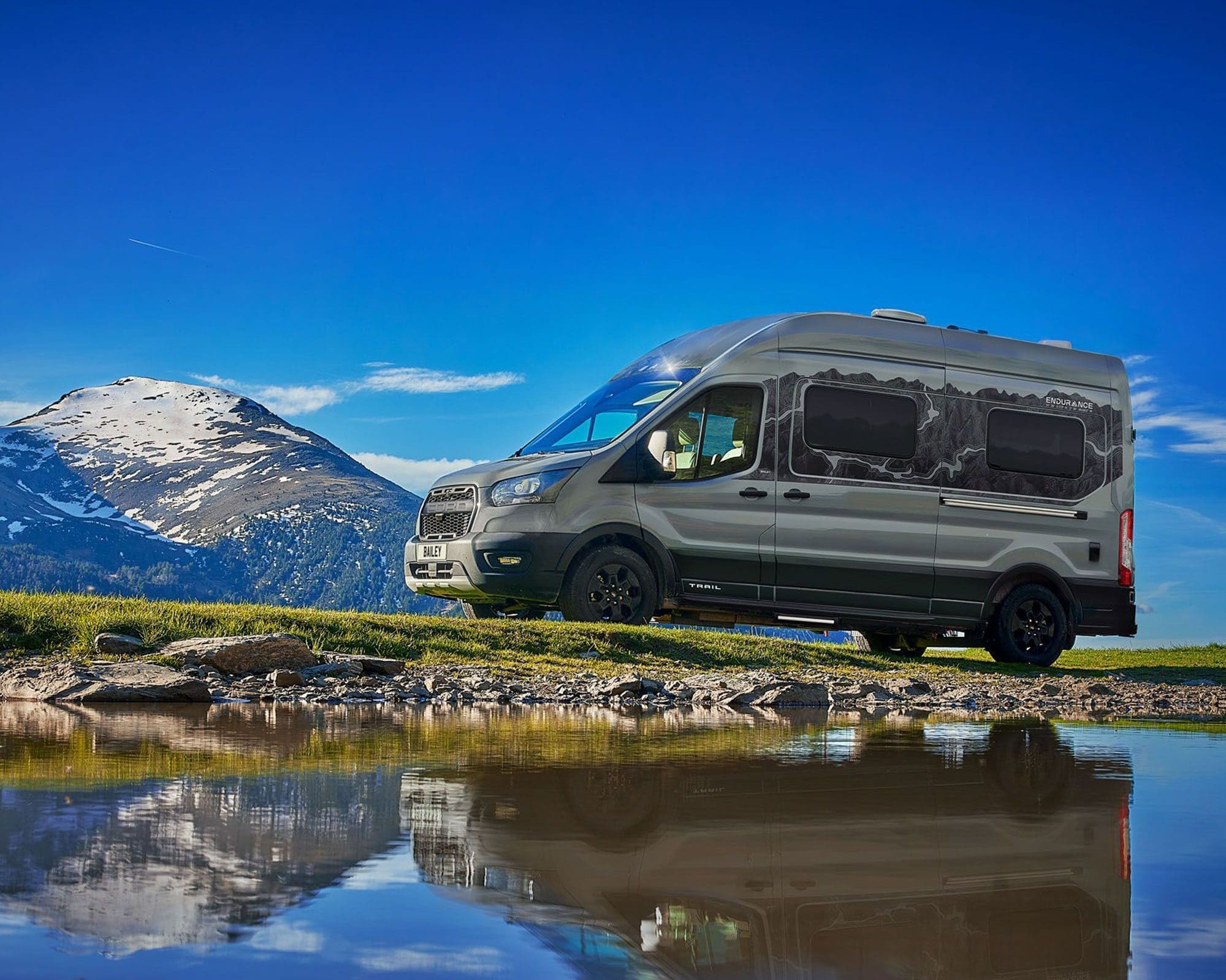 A sleek gray camper van parked beside a tranquil lake with mountainous scenery in the background.